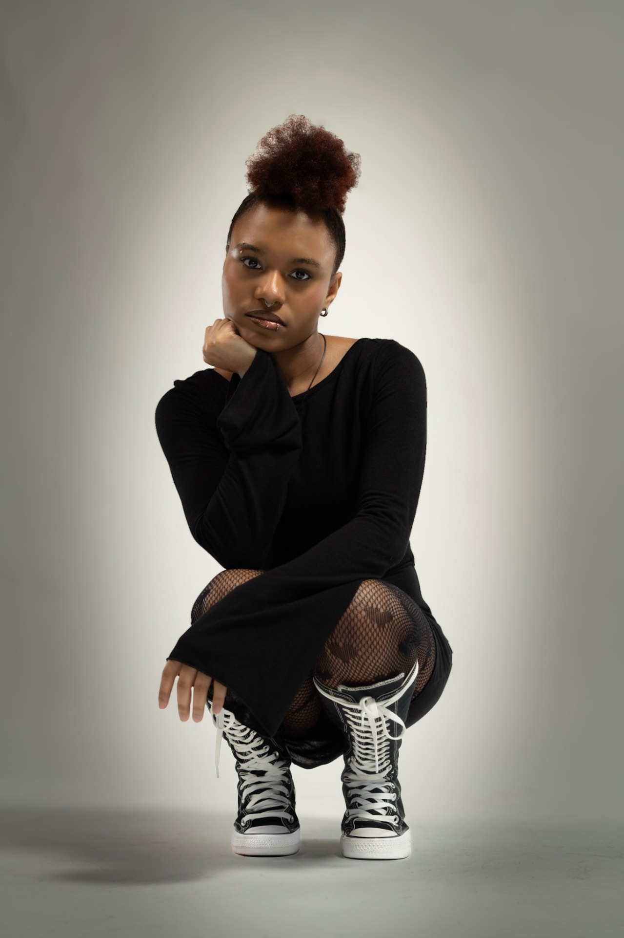 Studio portrait of a young woman in a black dress and high-top sneakers, crouched against a soft grey backdrop