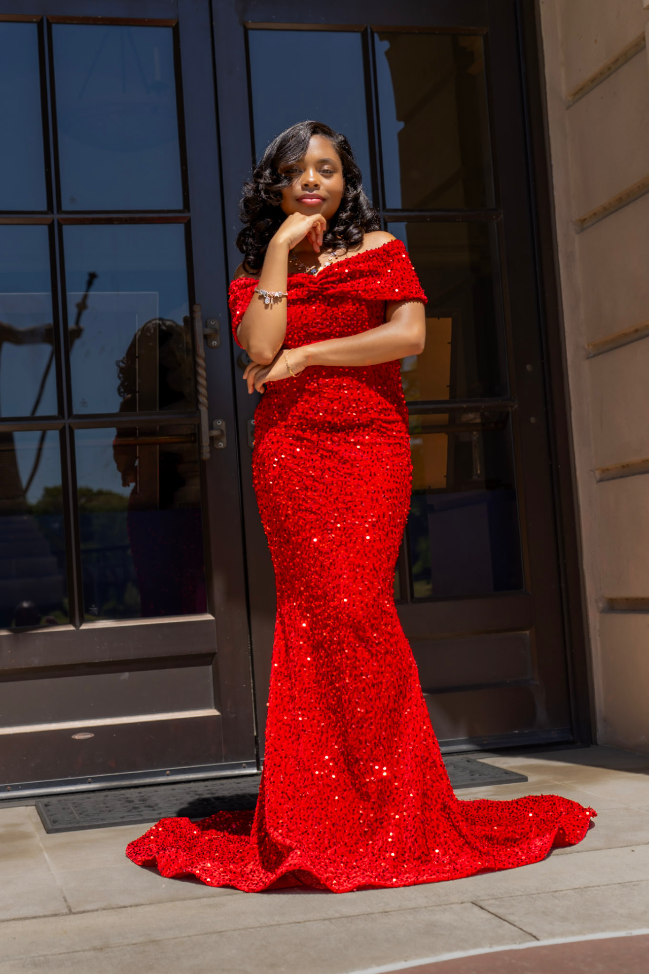 Portrait of a woman in a floor-length red sequin gown standing before tall dark glass doors in bright daylight